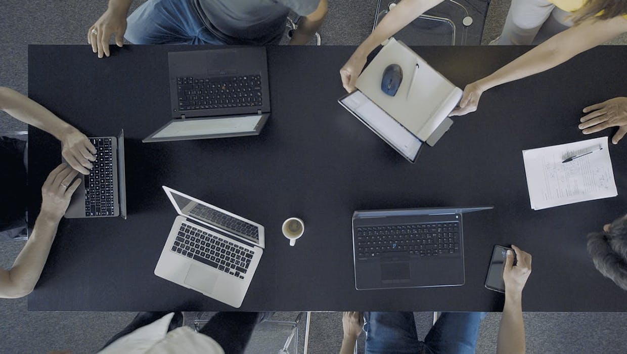 Vue aérienne d'ordinateurs portables posés sur une table autour de laquelle sont assises plusieurs personnes.