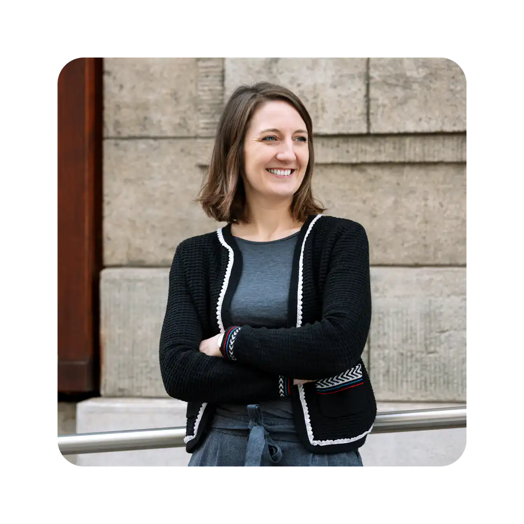 Femme souriante aux bras croisés, vêtue d'un pull noir, posant devant un mur de pierre.