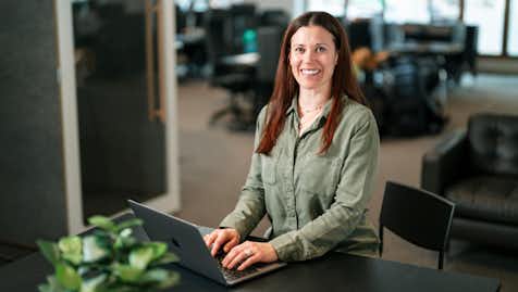 Smiling woman with long brown hair, wearing a green shirt, typing on a laptop at a desk in an office.