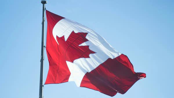 Red and white Canadian flag with a central red maple leaf, fluttering from a flagpole against a clear blue sky.