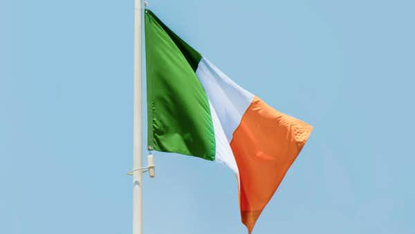 The green, white, and orange national flag of Ireland flies on a flagpole under a clear blue sky.