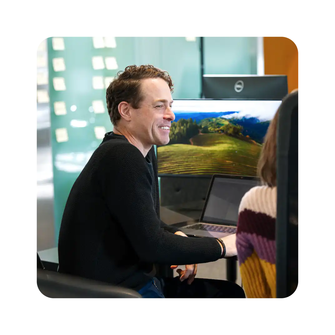 Eine Person mit lockigem Haar, die einen schwarzen Pullover trägt, lächelt an einem Laptop in einem modernen Arbeitsbereich mit einer grünen Landschaft auf dem Bildschirm.