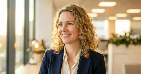 Smiling woman with curly hair, wearing a navy blazer and white blouse, stands near large windows in an office setting.