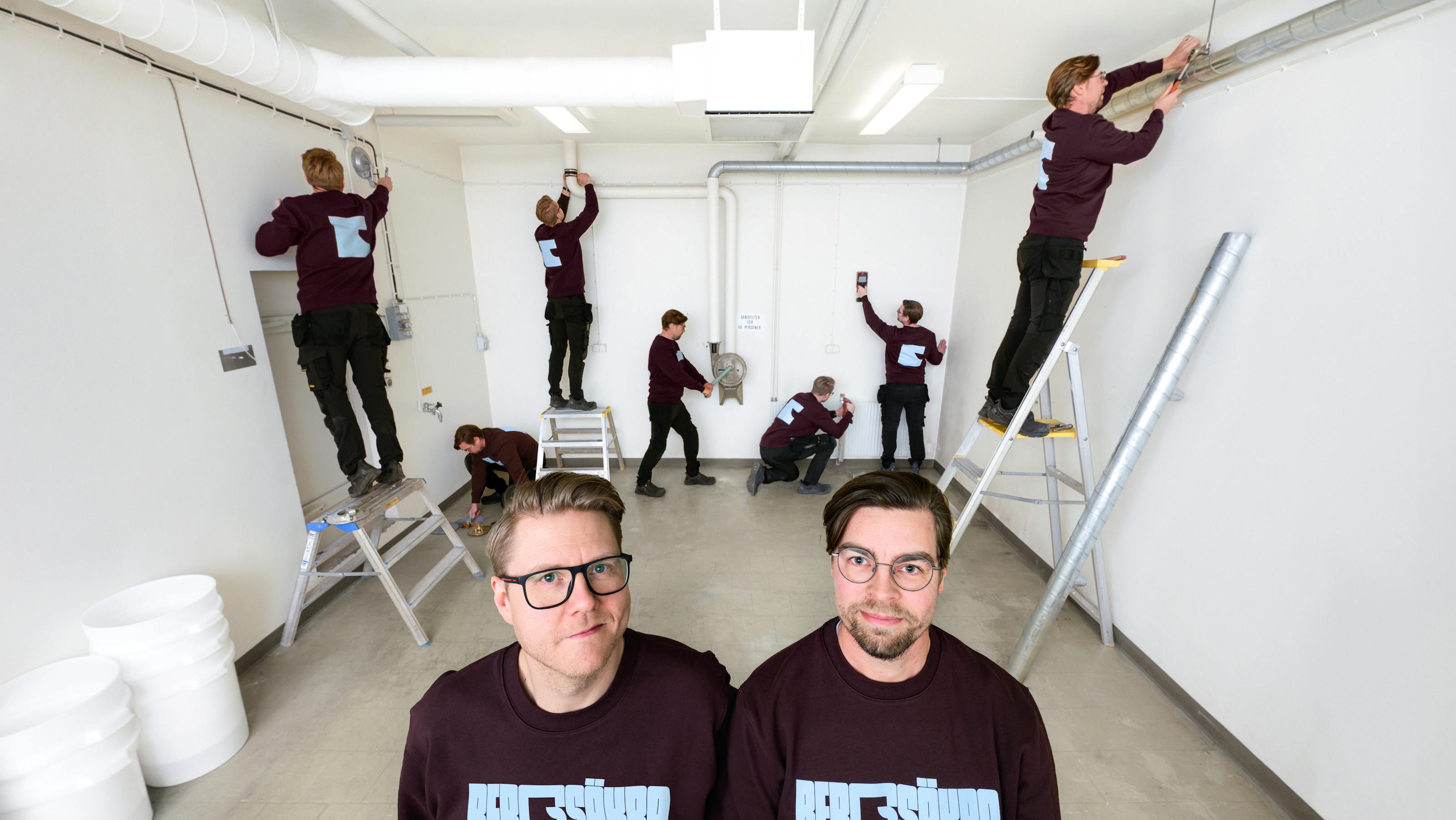 Två st skyddsrumstekniker i förgrund med flera kopior av sig själv som utför arbeten i bakgrunden
