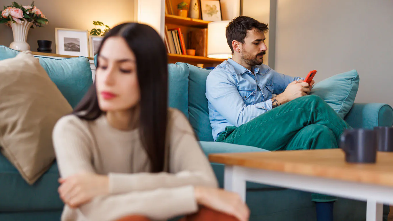 A couple at home; the woman looks pensive while the man casually browses his phone