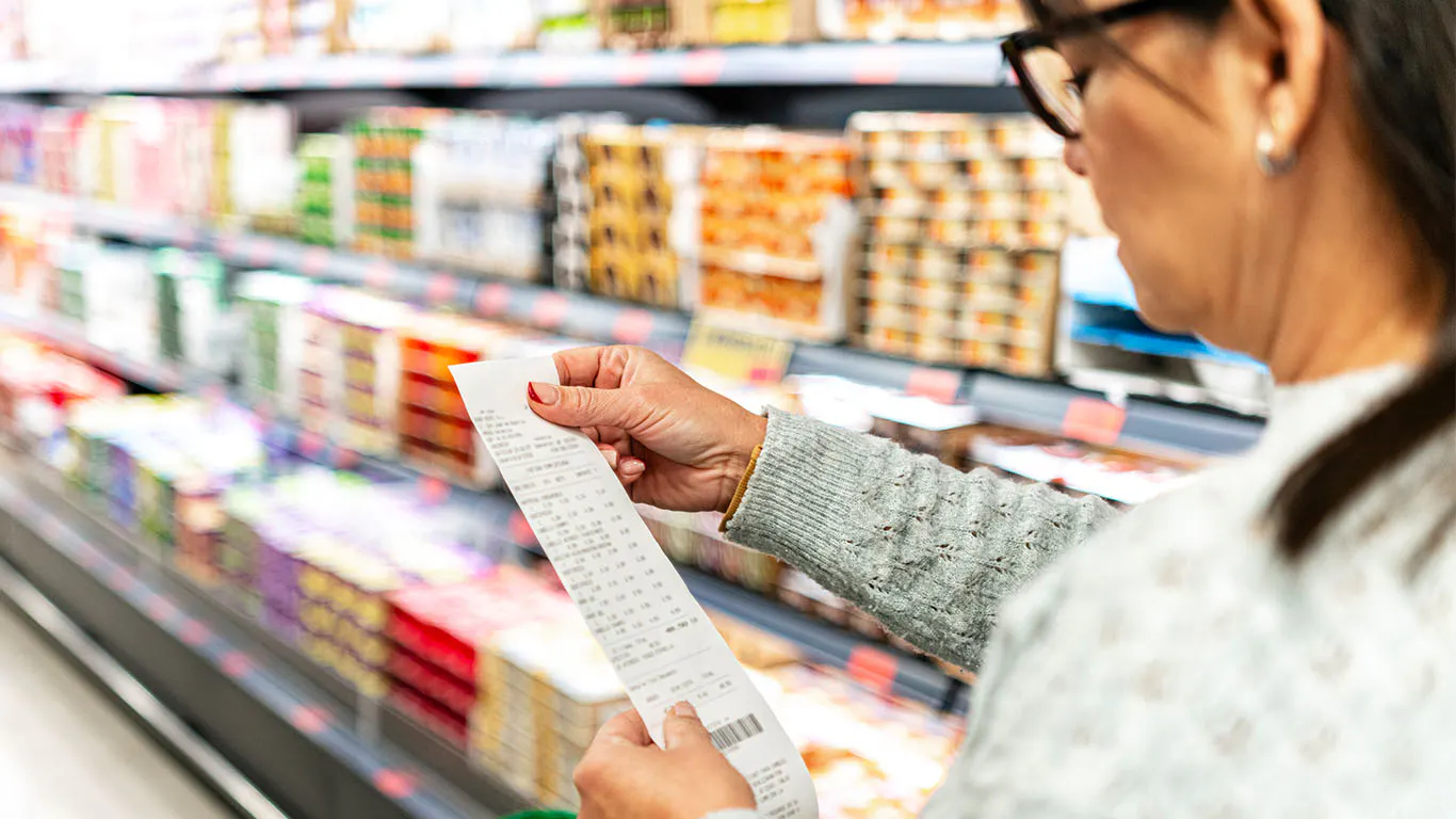 A middle-aged woman standing in a grocery aisle looking at a long receipt