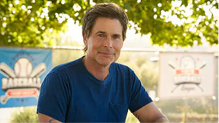 Rob Lowe in a navy T-shirt standing outdoors in front of trees and baseball banners