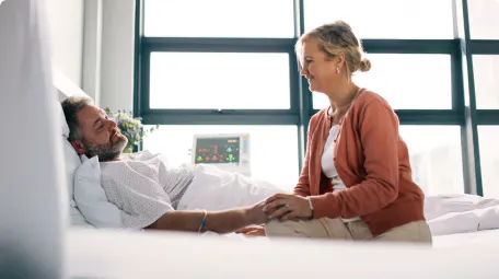 A woman in a coral cardigan smiling and holding the hand of a man lying in a hospital bed