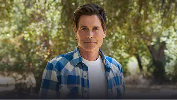 Rob Lowe in a blue plaid shirt over a white T-shirt, standing among trees
