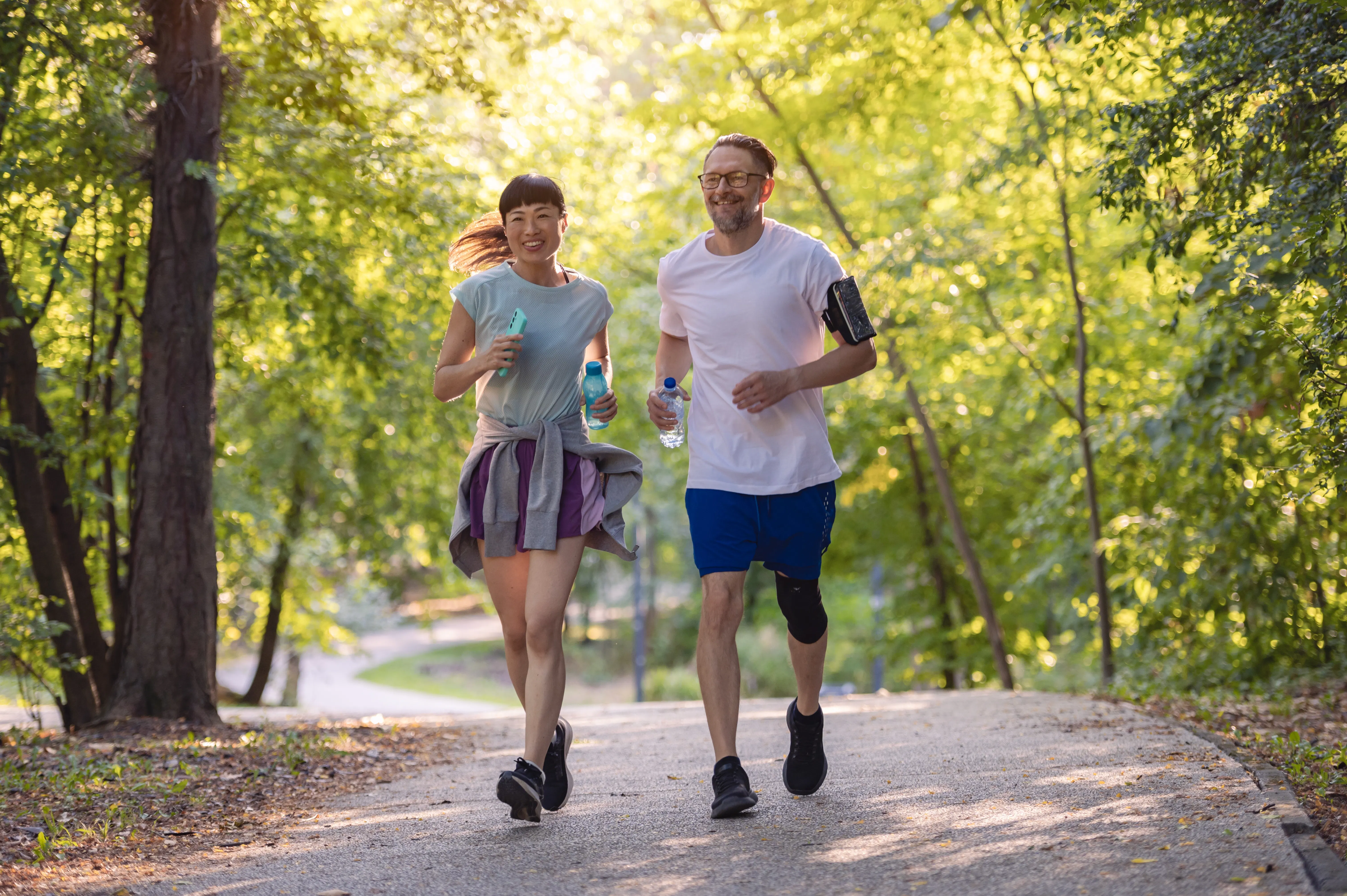 Two people jogging along a tree-lined park path, carrying water bottles on a sunny day.