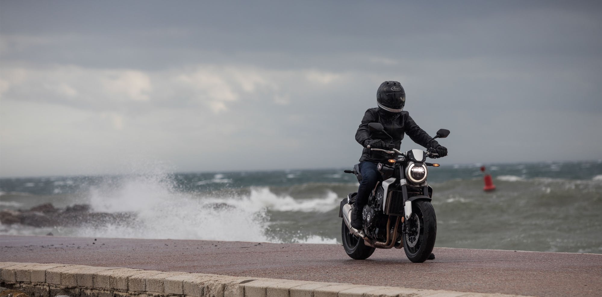 Honda CB1000R in graphite black colour, being ridden on the street road by the bay