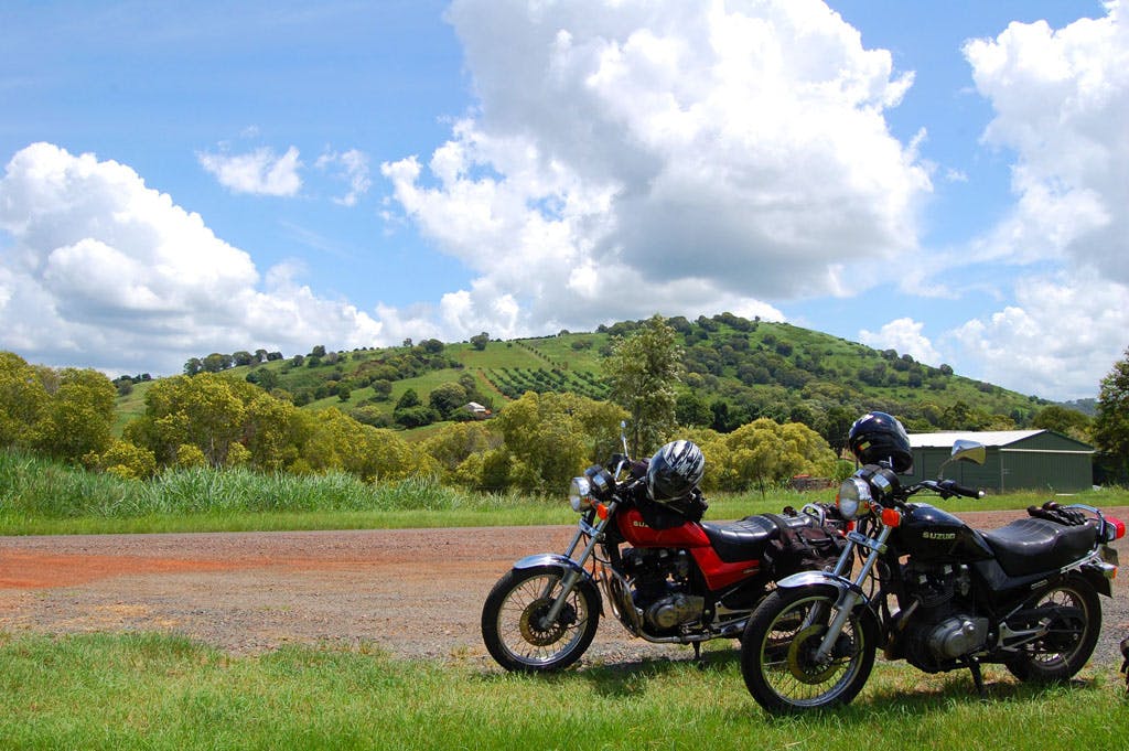 a black and a red motorcycle near a dirt road on a sunny day with clean skies and white clouds
