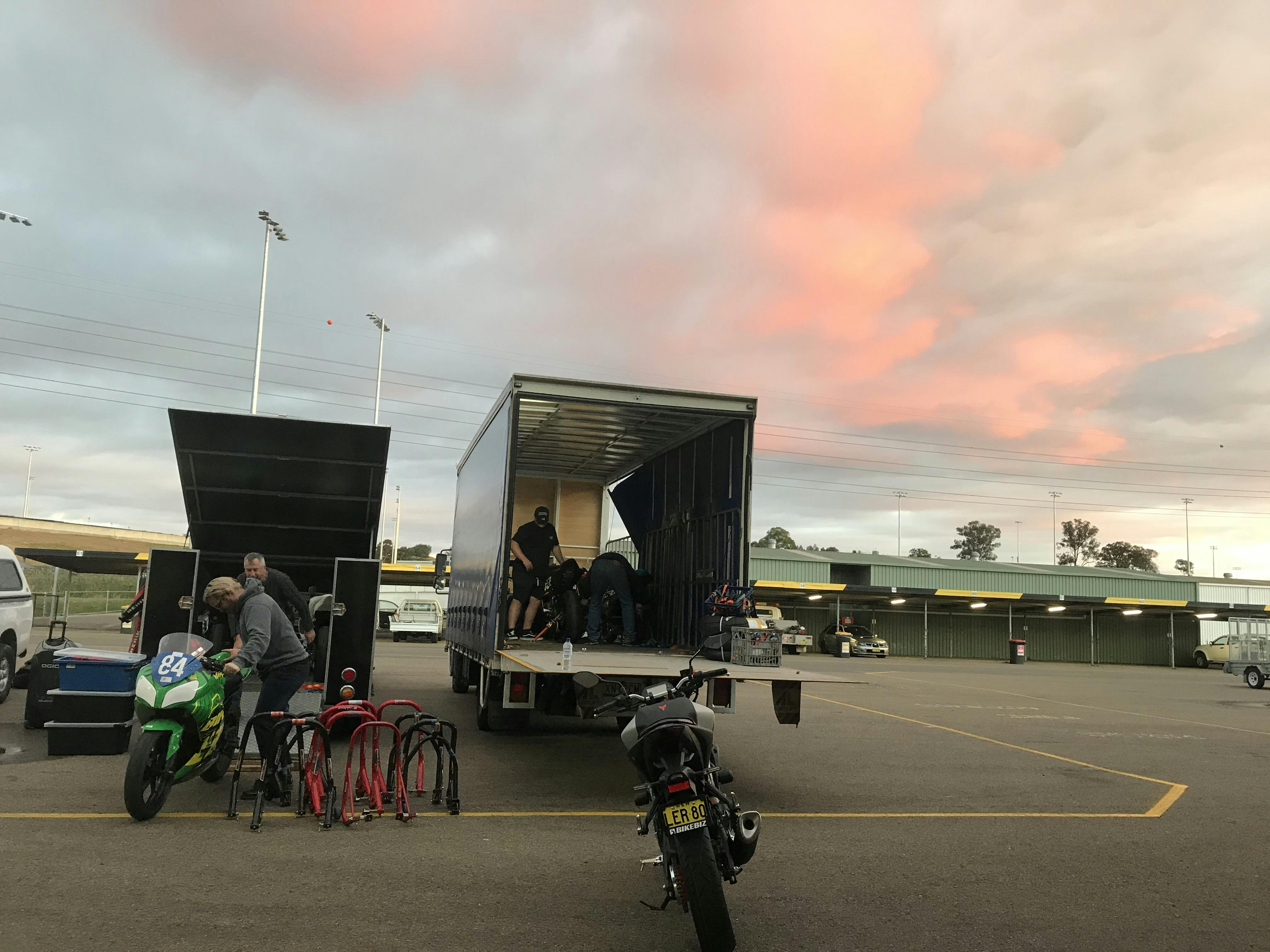 Motorcycles being loaded into a truck with a pink and blue sky background
