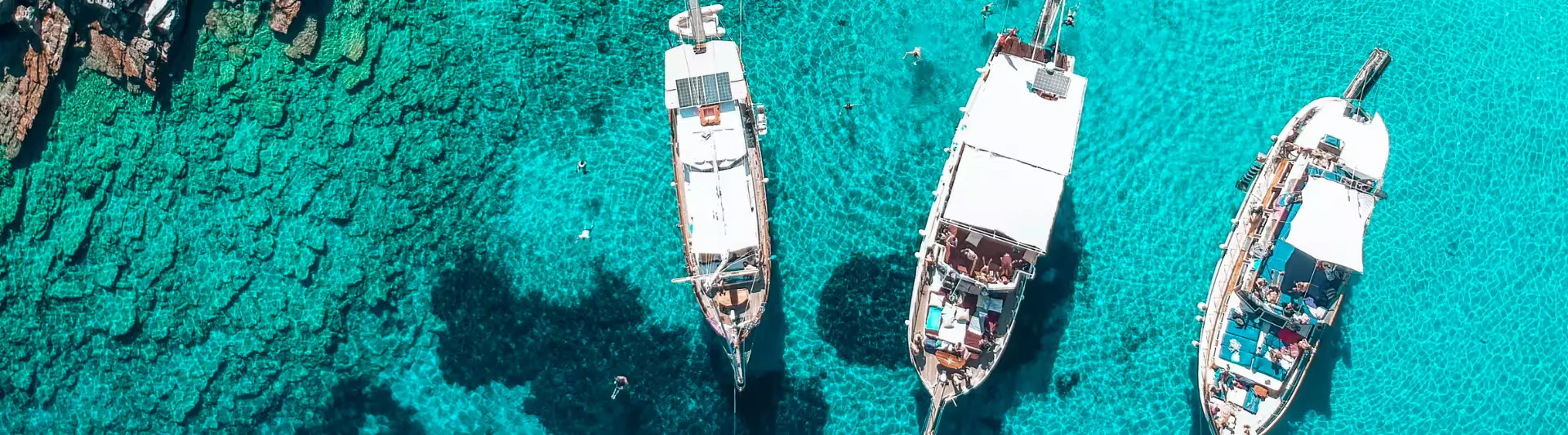 Bodrum açıklarında Ege Denizi'nin kristal berraklığındaki turkuaz sularında demirlemiş üç adet beyaz tekneye tepeden bir bakış. Dinlendirici ve güneşli kıyı manzarası, biletiniz.com'un İstanbul-Bodrum uçak bileti seyahat rehberine huzurlu bir giriş sunuyor.