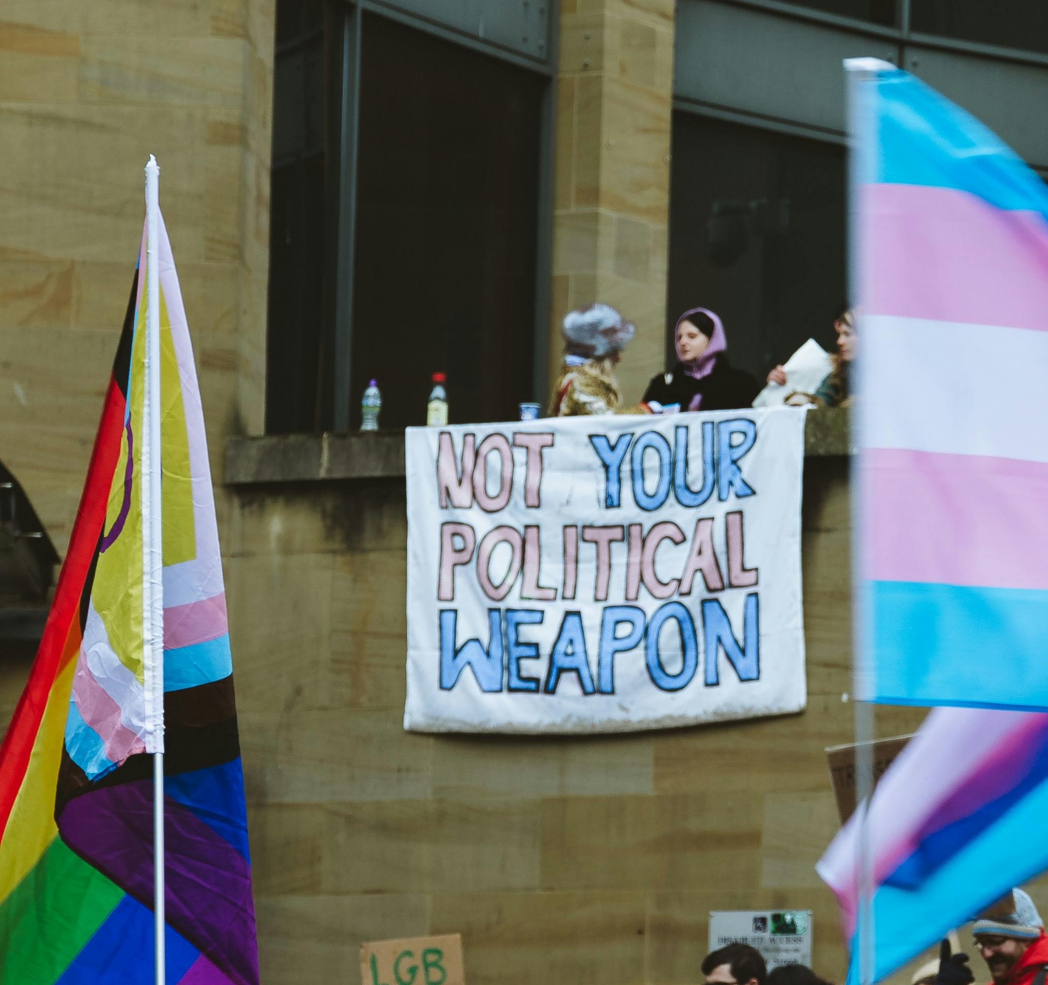 People holding sign saying "Not Your Political Weapon" with Trans and LGBT flags. 
Photo by Thiago Rocha on Unsplash

