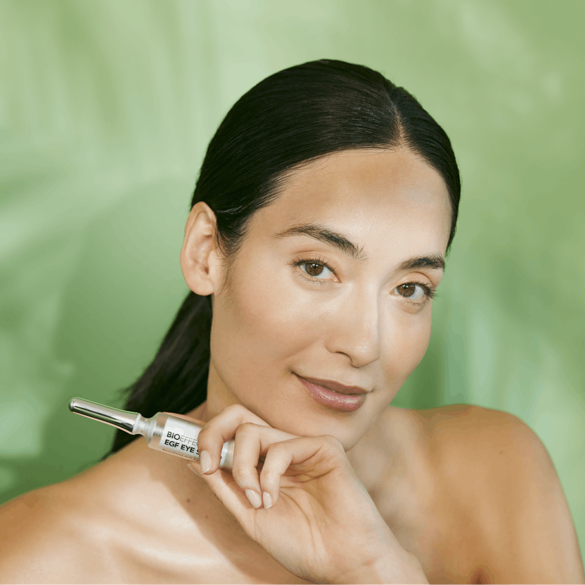 Woman with glowing skin holding Bioeffect EGF Eye Serum against a green background.