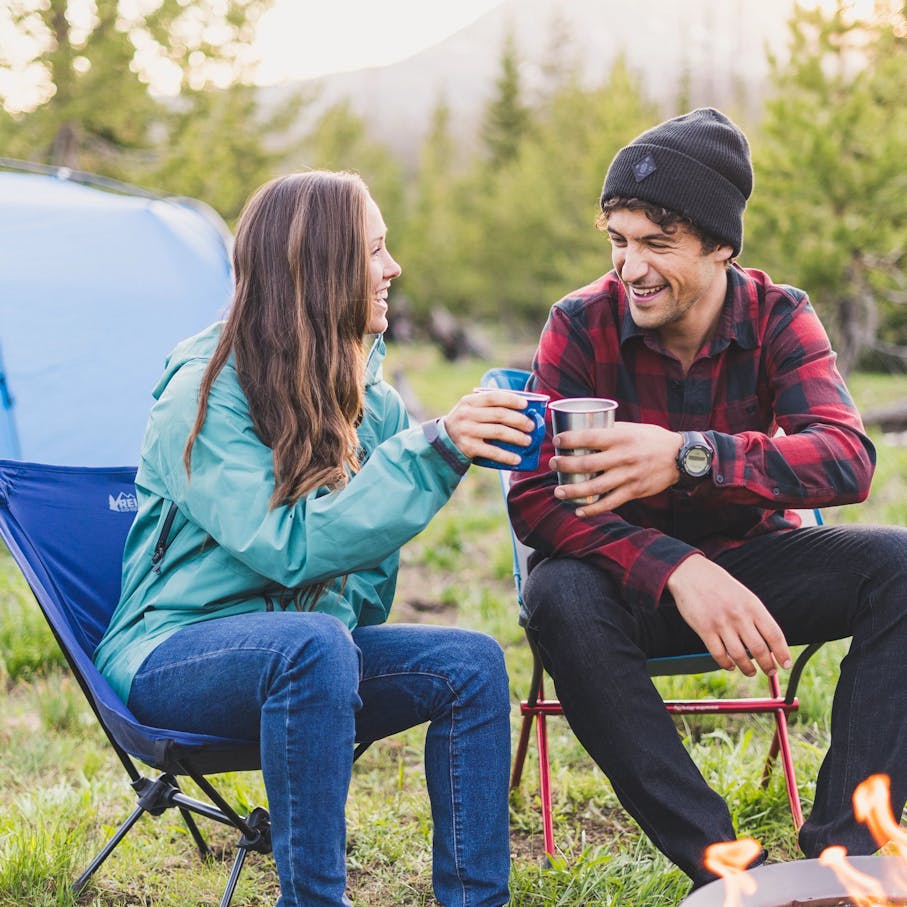 Man and woman sitting in camping chairs in front of a campfire in BD apparel with cups in their hands