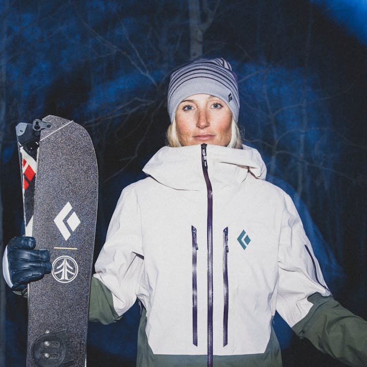 A splitboarder stand ready for her tour in Black Diamond Recon LT Shell. The mountains behind her are barely illuminated in the predawn glow. 
