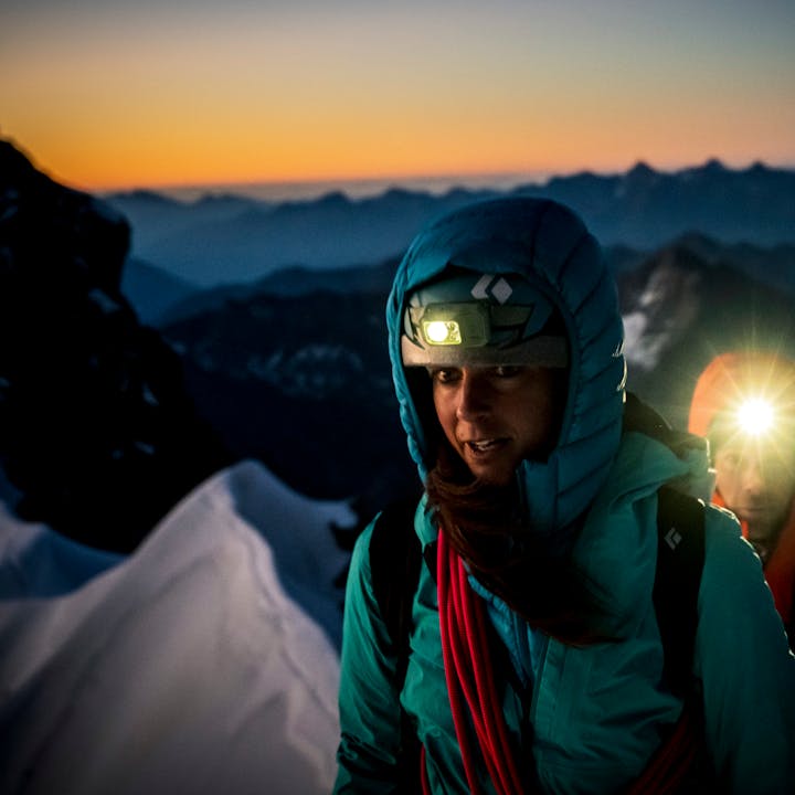 Two alpinists look down their route at dawn bundled up and wearing Black Diamond headlamps. 