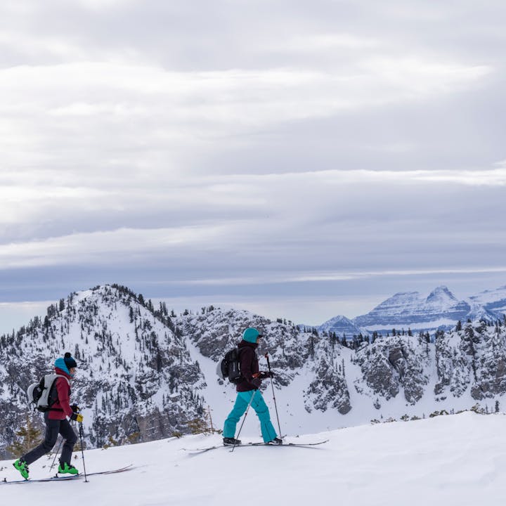 Two skiers walk along a snowy ridgeline, the sky cloudy behind them. 