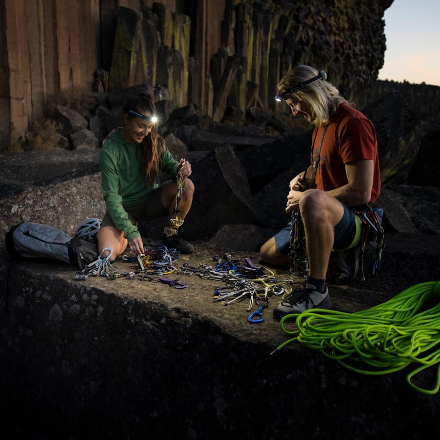 Two climbers organizing their rack in front of a climbing wall using the light of BD headlamps at dawn