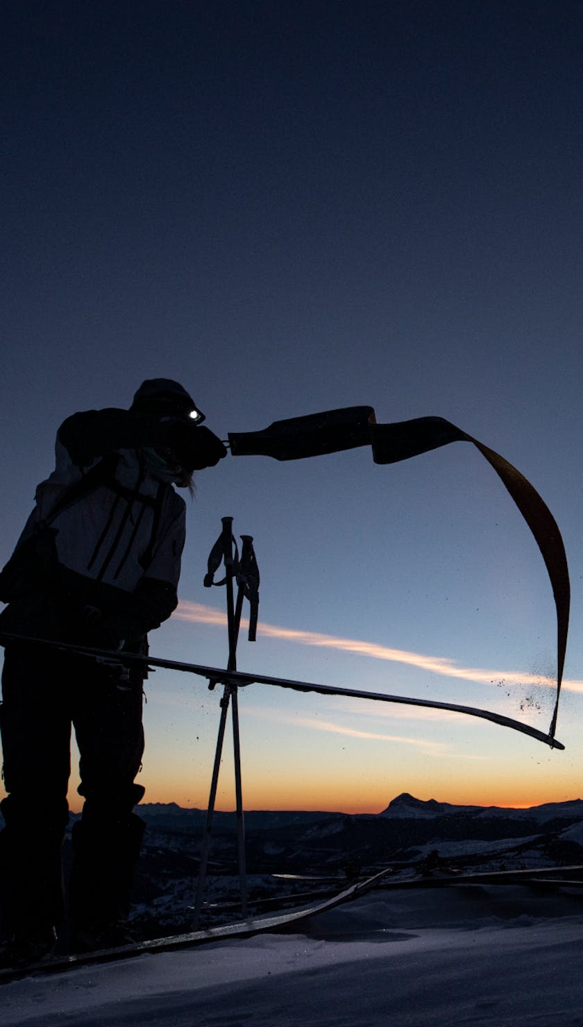 A skier rips skins and transitions from tour to ski mode at the top of a mountain by headlamp. 
