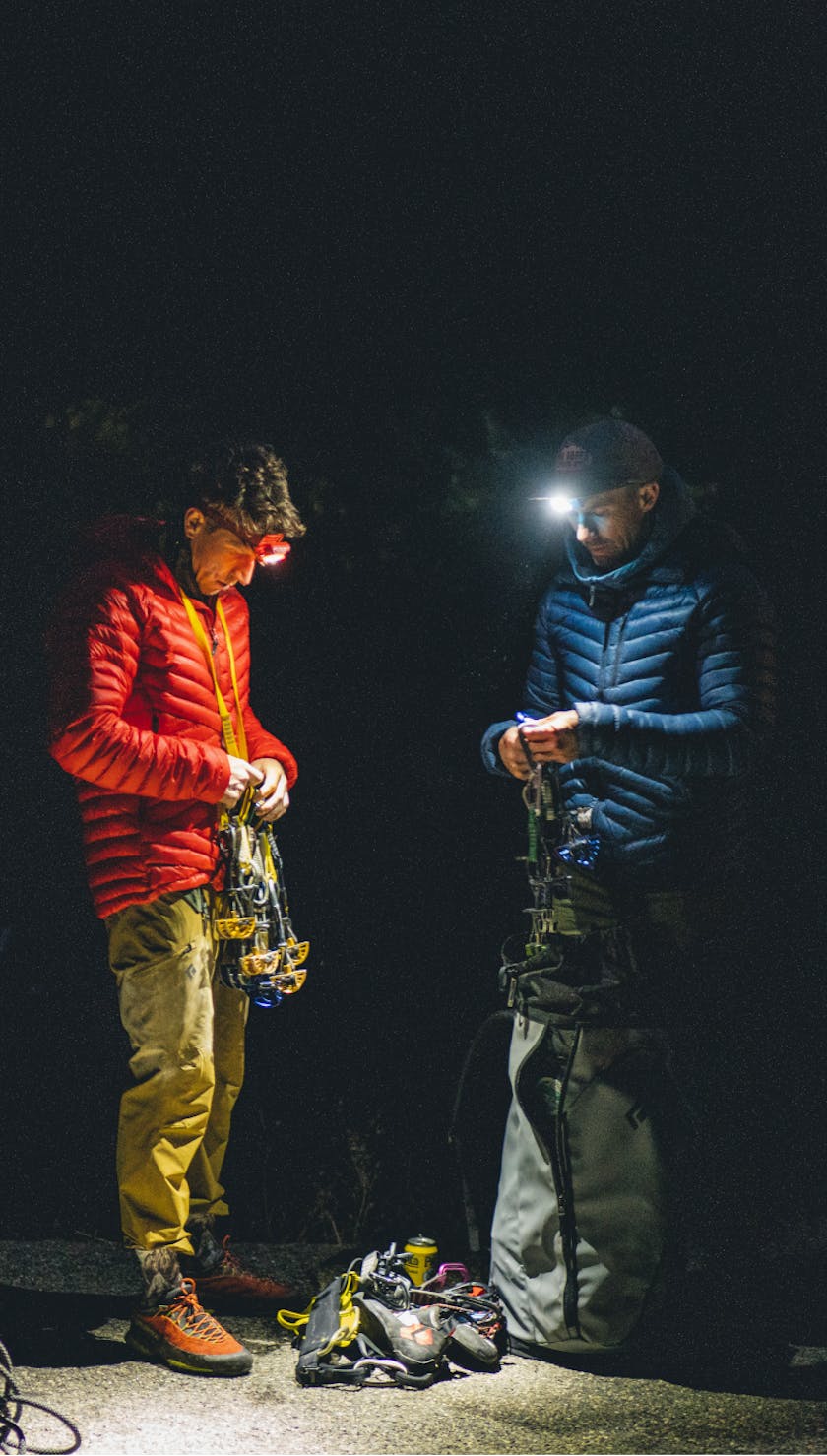 Two climbers sort their gear by headlamp wearing the Black Diamond Approach Down hoody. 