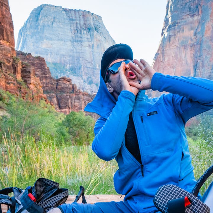 A climber wearing a Black Diamond Hoody cheers up towards the wall with the epic scenery of Zion behind him. 