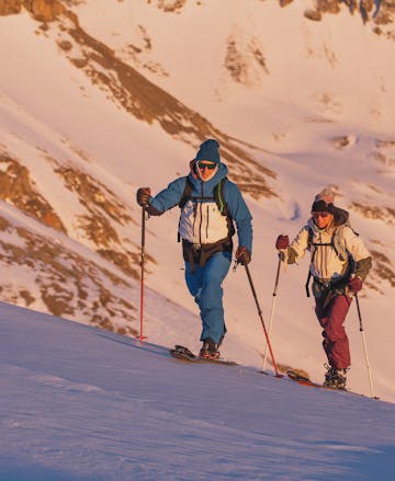 Deux skieurs partent en patrouille à l'aube, dévalant la montagne sous le soleil matinal portant des vêtements Black Diamond.