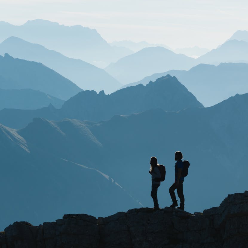 Two hikers stand on a ridgeline looking out at an expansive mountain range. 