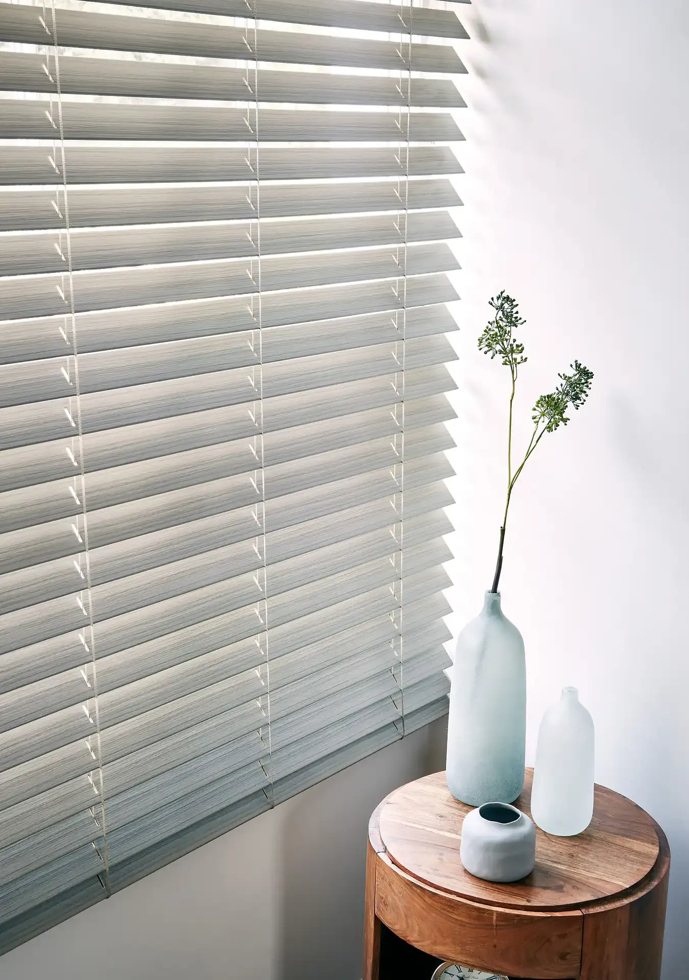 Horizontal plaswood blinds filtering daylight across a corner with a round wooden side table holding frosted vases and a tall green sprig against a pale wall.