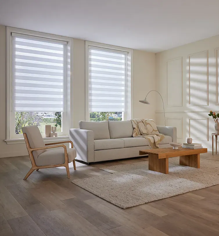 Double roller blinds filtering soft daylight across a neutral living room with a grey sofa, wooden armchair, textured rug and low wooden coffee table.