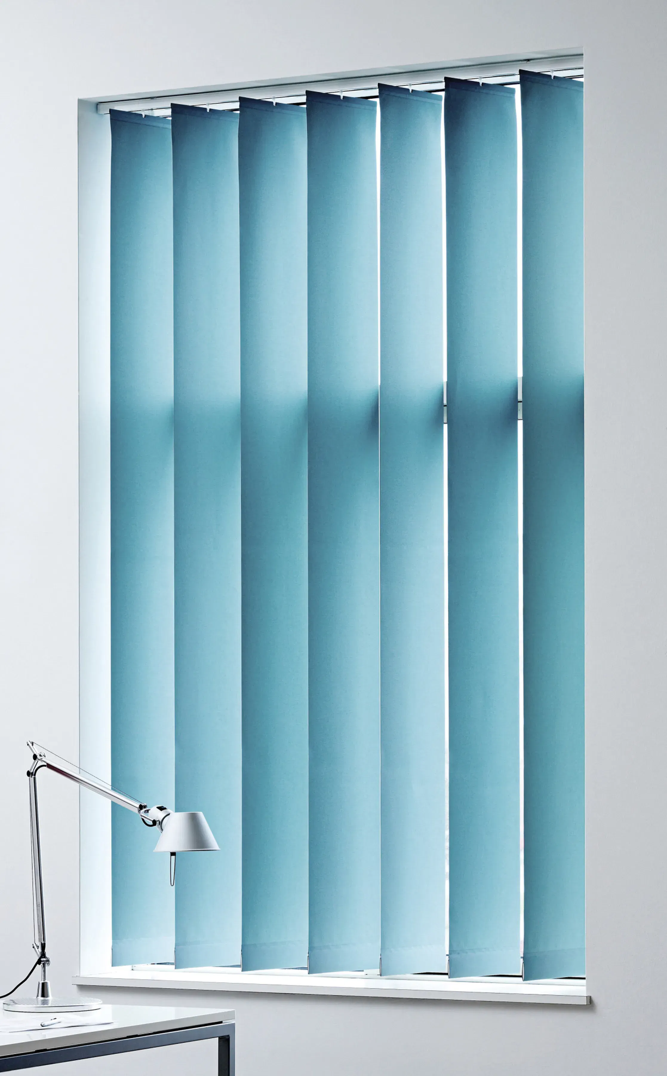 Vertical blue blinds covering a tall window, slightly tilted to let thin shafts of light through, in a minimal white office setting beside a chrome desk lamp.
