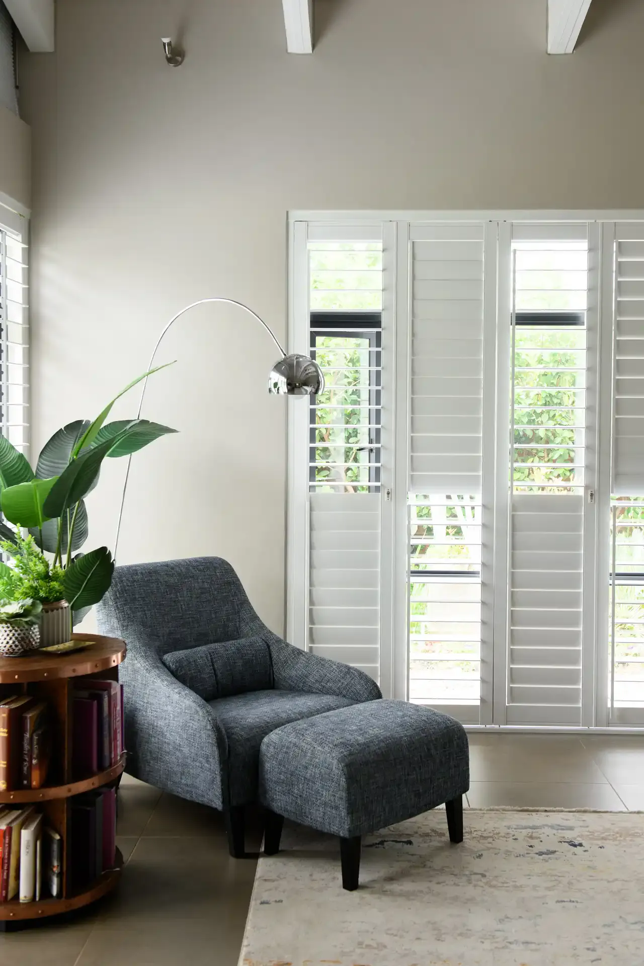 White plantation shutters filtering daylight across a cozy living room with a blue armchair and ottoman, arc floor lamp and potted plant.