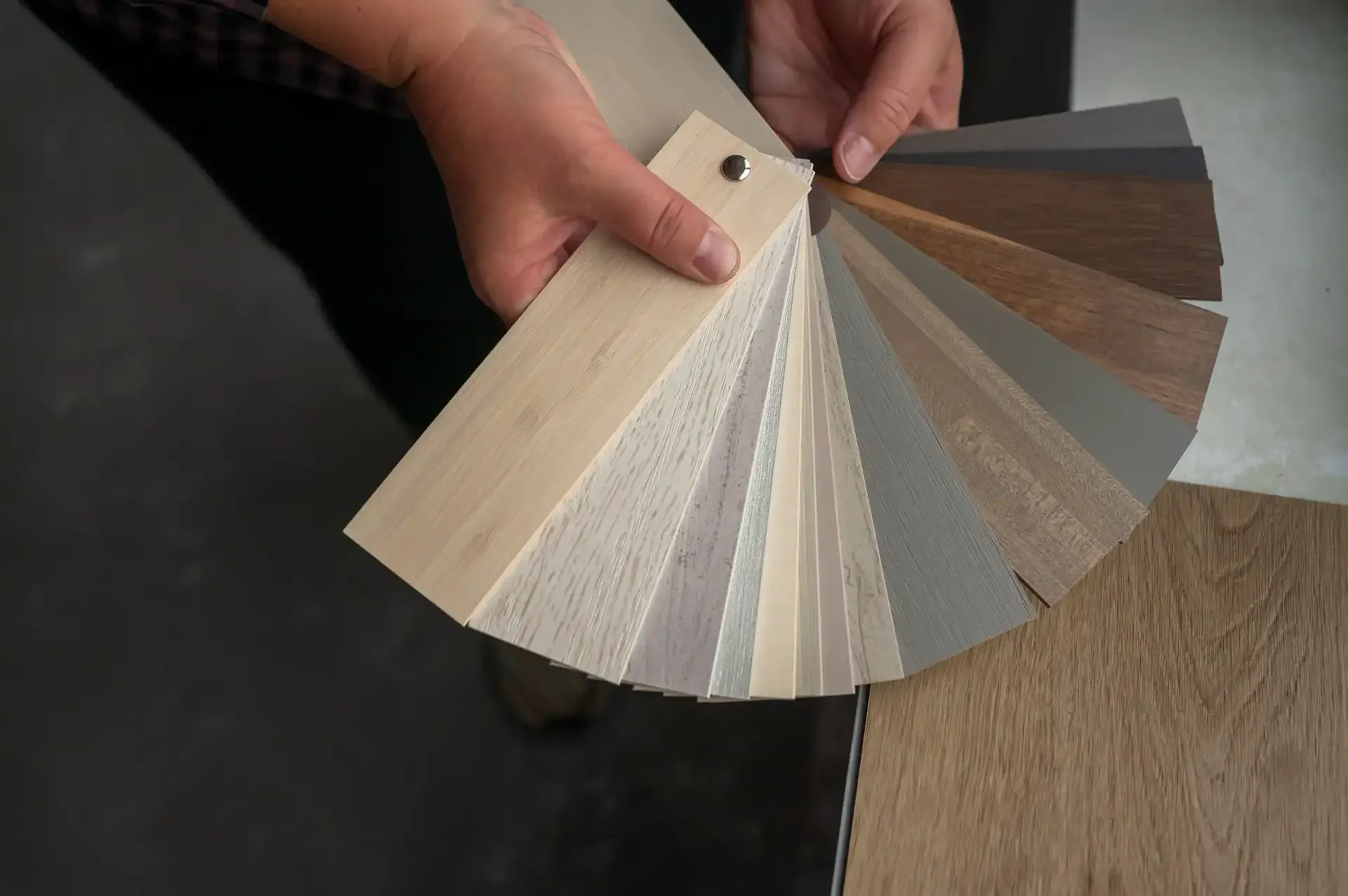 Wood-effect blind slat samples fanned in hands, being examined over a light wood tabletop in an interior showroom or workshop.