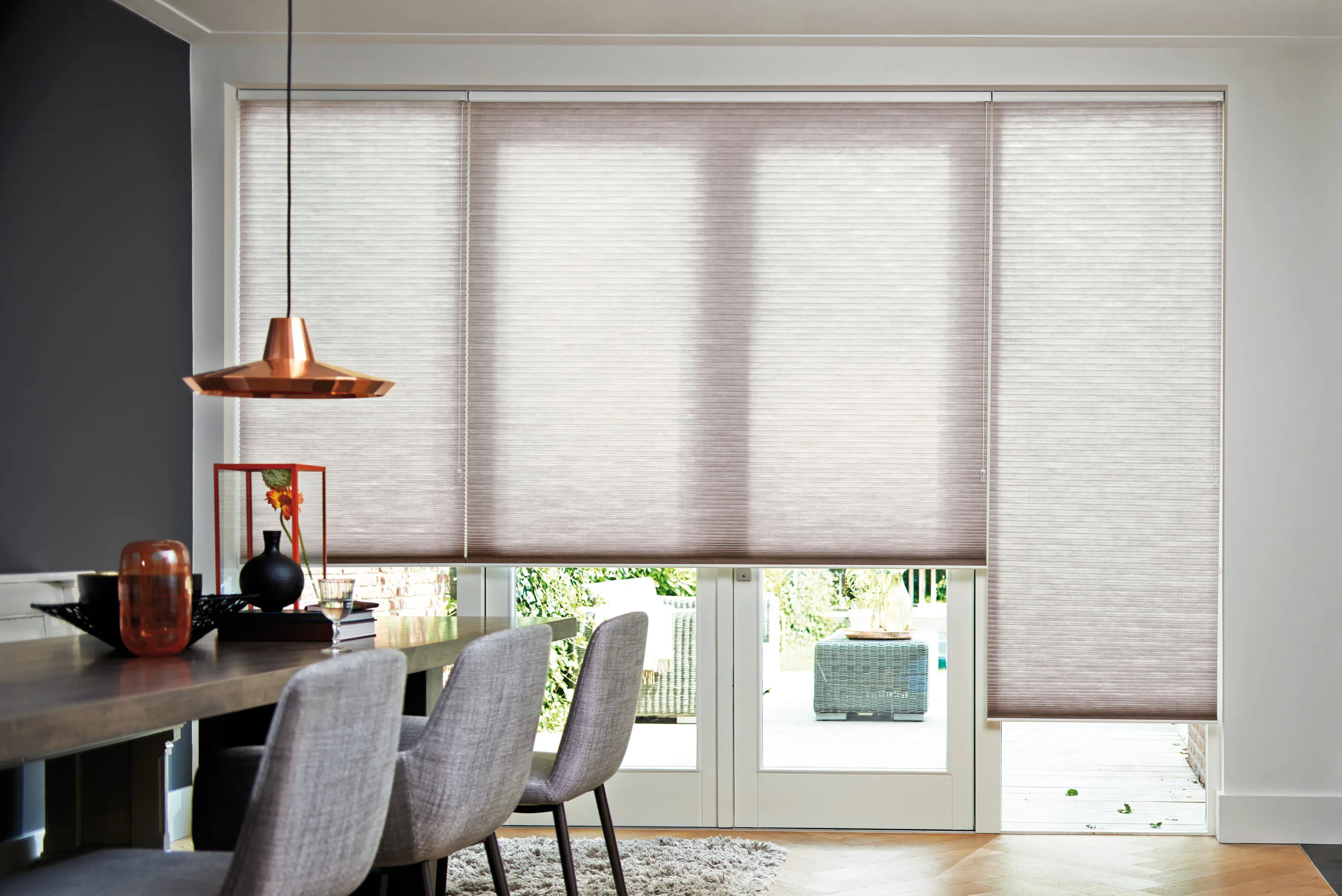 Neutral honeycomb blinds covering patio doors, diffusing light; dining table with grey upholstered chairs and copper pendant lamp in a modern open-plan dining