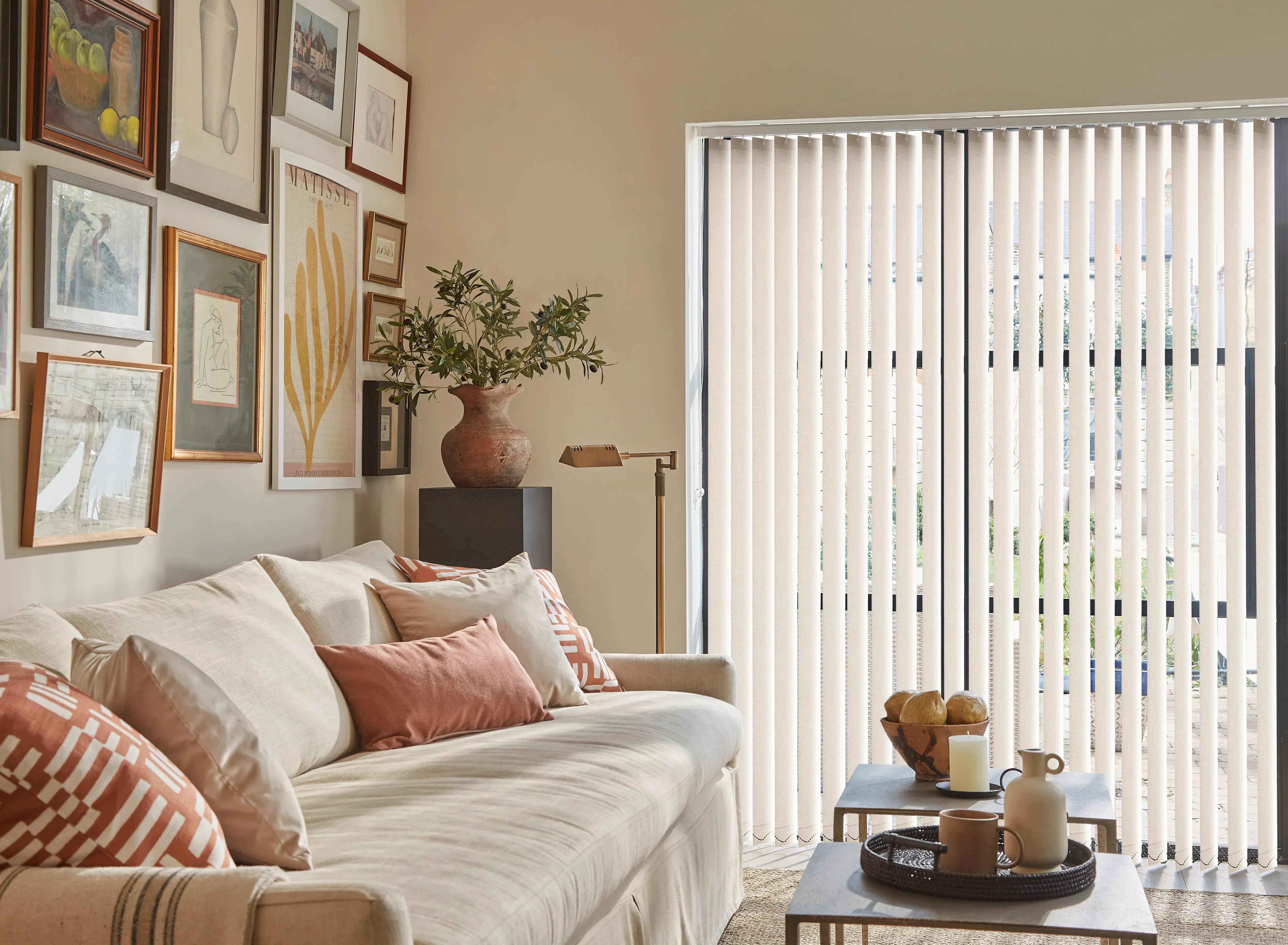 Vertical blinds filtering daylight across a cozy living room with beige sofa, terracotta cushions, gallery wall (text: MATISSE) and a coffee table with ceramics
