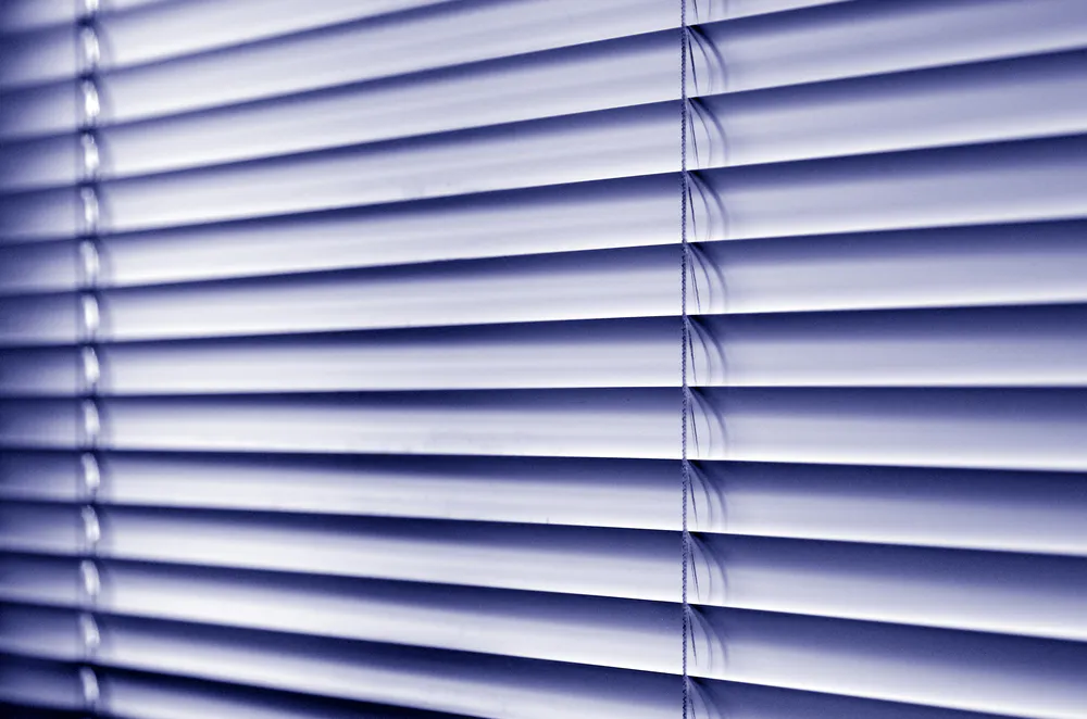 Horizontal venetian blinds tilted closed, casting soft striped shadows across an interior window, cords visible and cool blue-toned light subtly filtering
