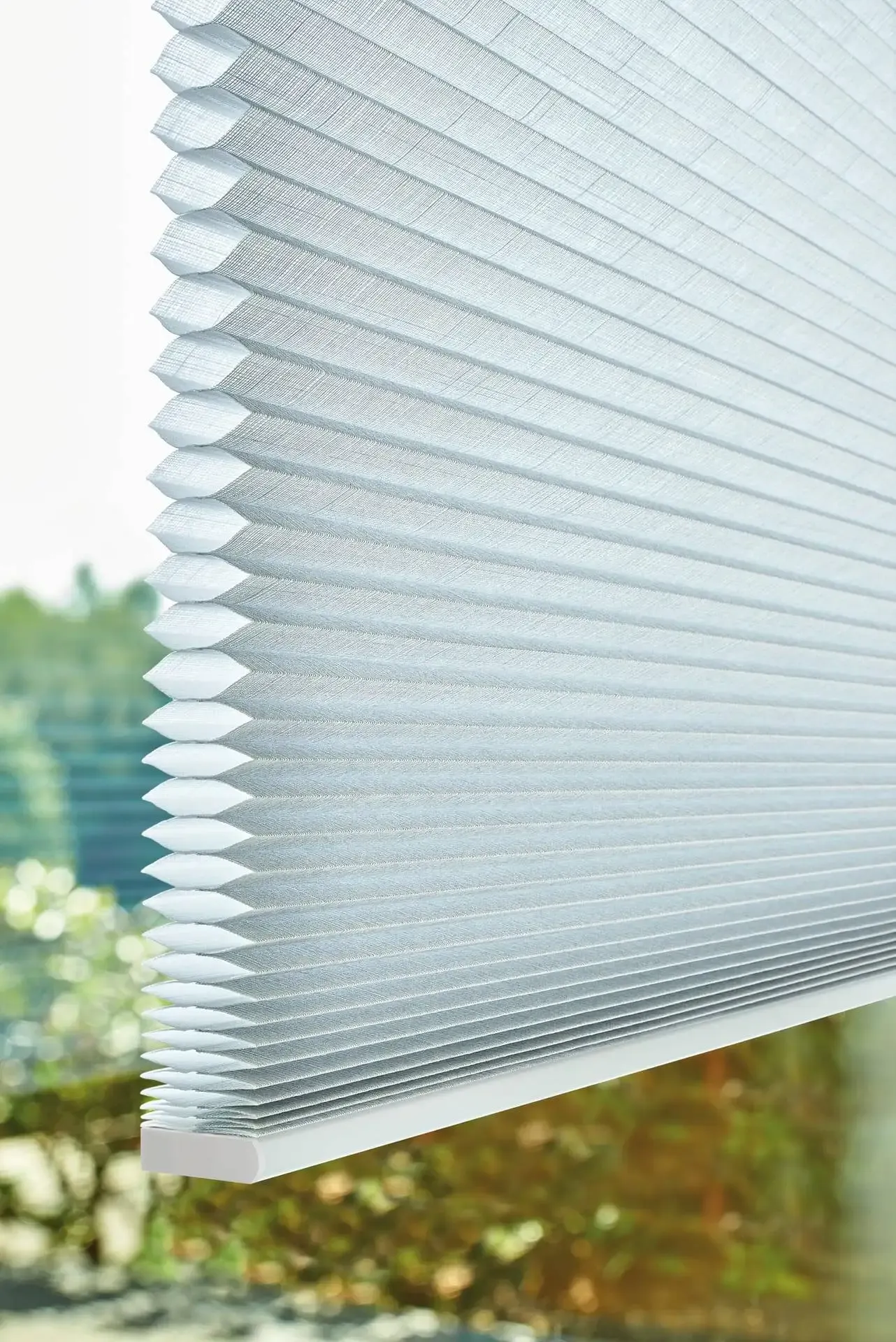 Pleated cellular blind hangs partially lowered, its honeycomb folds filtering daylight against a blurred green garden visible outside the window.