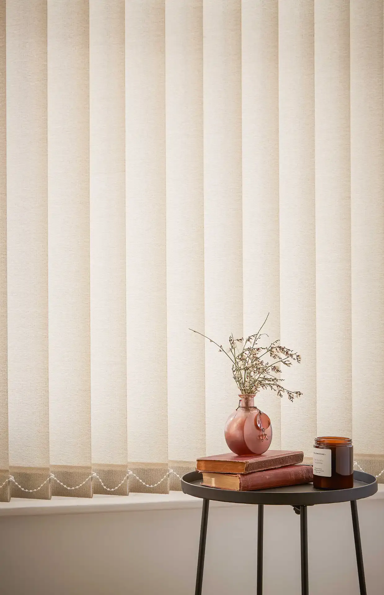 Beige vertical blinds covering a window, filtering light; in front a round side table holds a terracotta vase with dried stems, stacked books and a brown candle