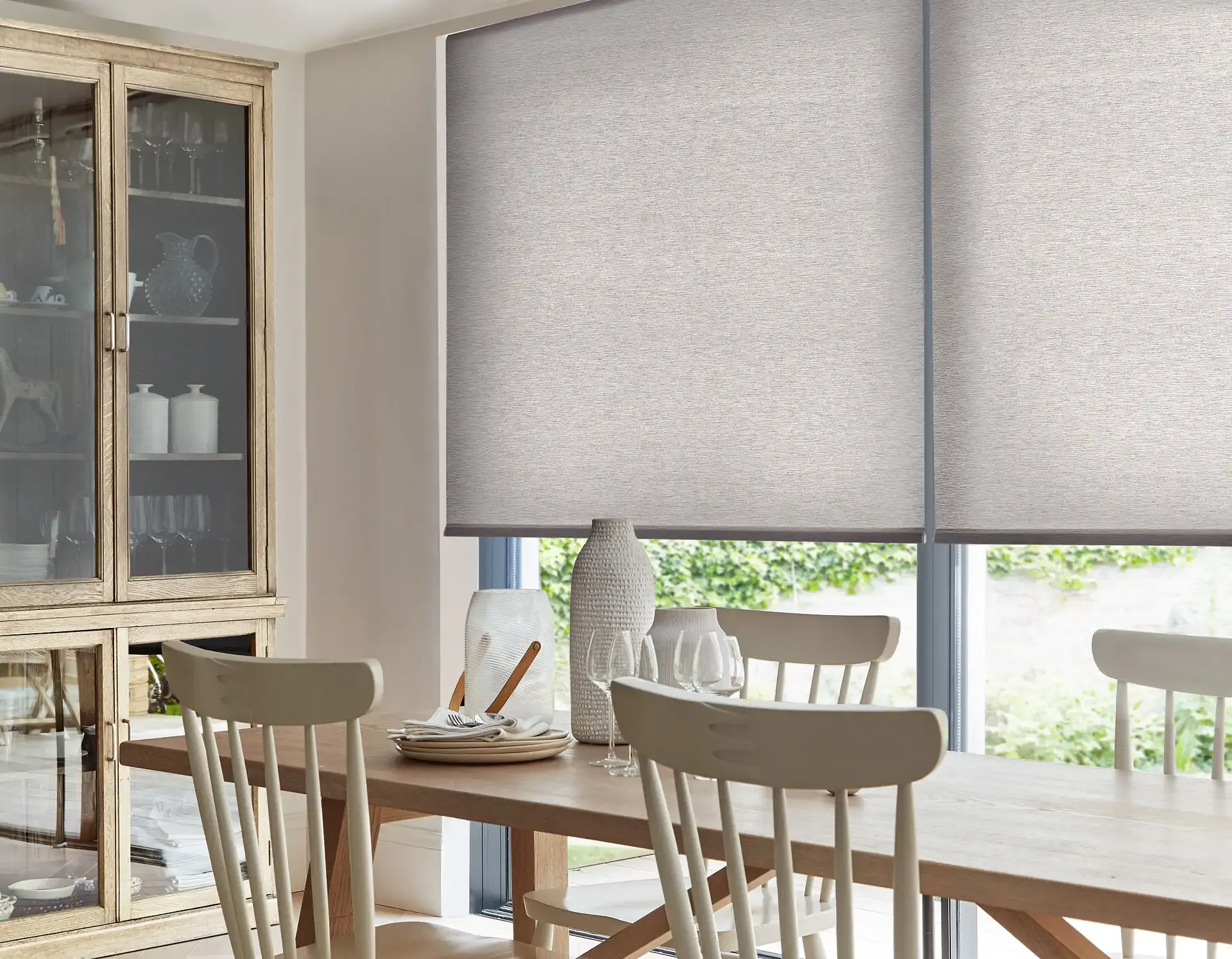 Neutral roller blinds filter light over a dining table set with plates and glassware, framed by cream chairs and a wooden glass-front cabinet in a bright dining