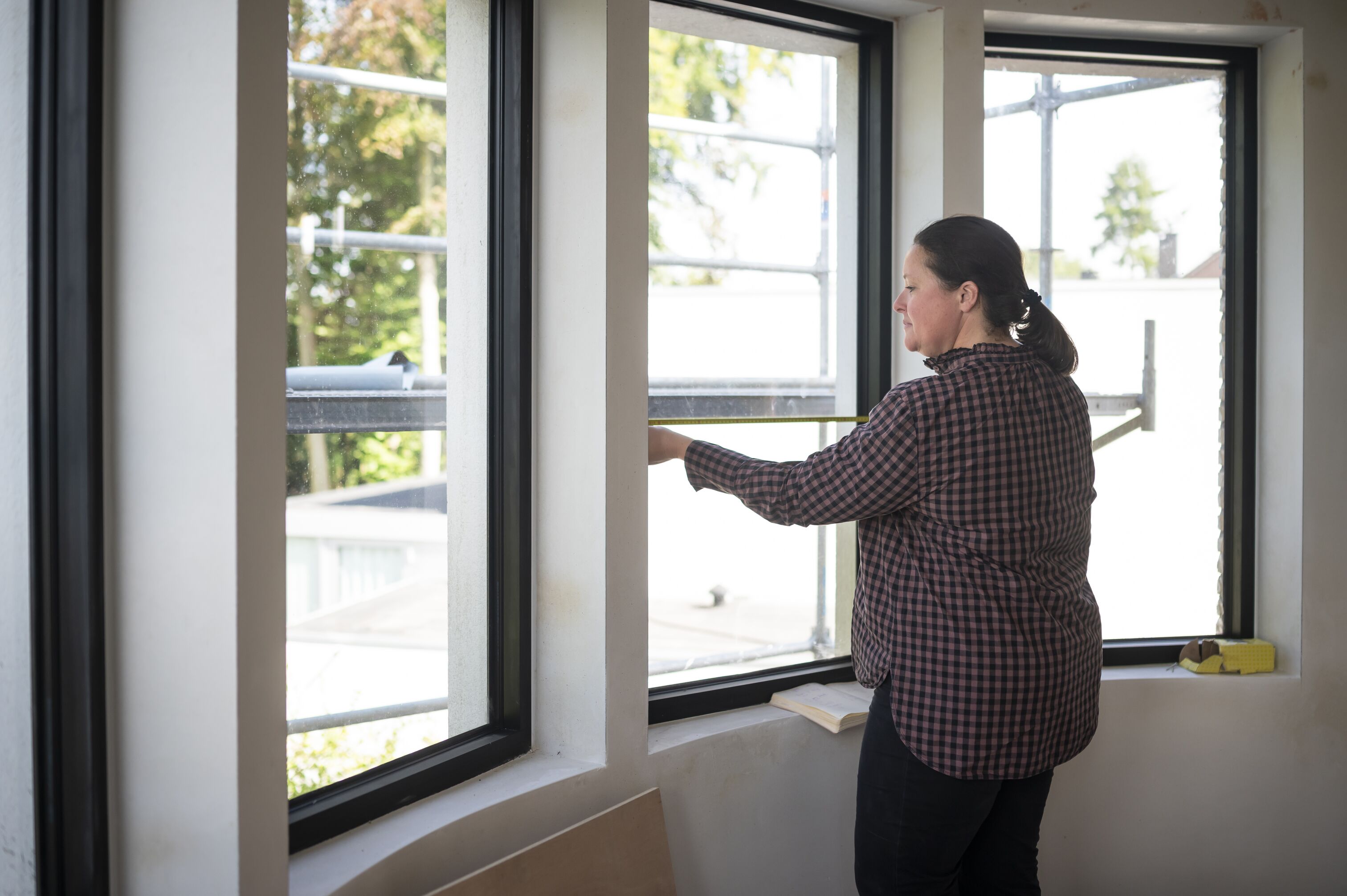 Window being measured by a woman with a tape measure inside a bright room, black-framed windows overlooking scaffolding and trees outside