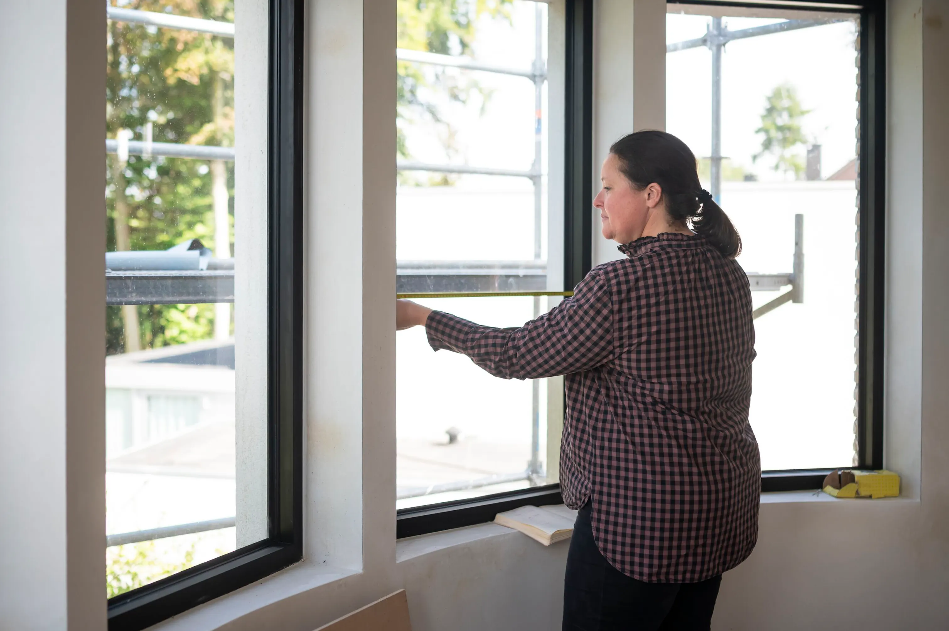 Window being measured by a woman with a tape measure inside a bright room, black-framed windows overlooking scaffolding and trees outside