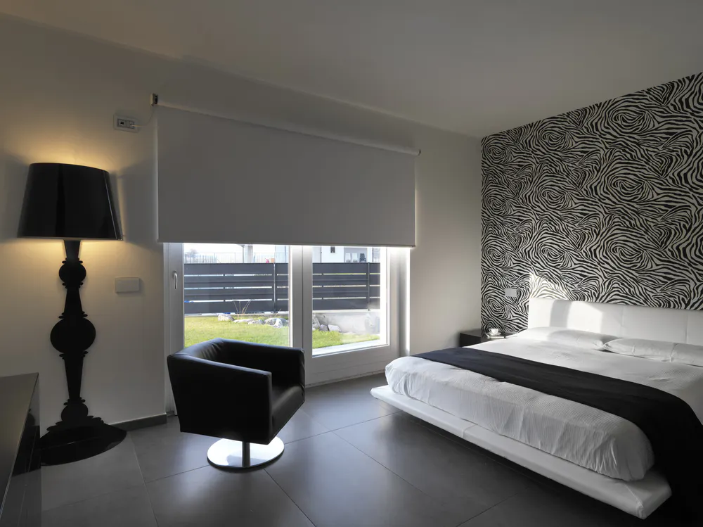Roller blind lowered over patio window in modern bedroom, blocking light; room features zebra-patterned accent wall, low white bed, black armchair and tall