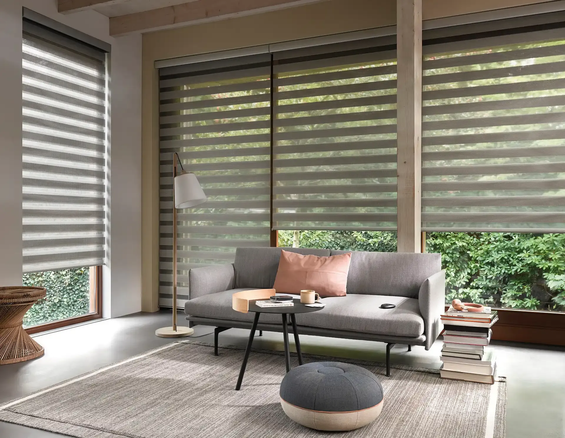 Double roller blinds filtering striped daylight across a cosy living room with grey sofa, floor lamp, pouffe and stacked books, framed by a leafy garden outside