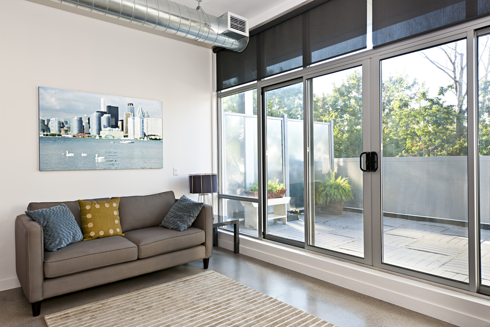 Roller blinds partially lowered over large sliding glass doors, filtering sunlight into a modern living room with a grey sofa, patterned cushions, cityscape