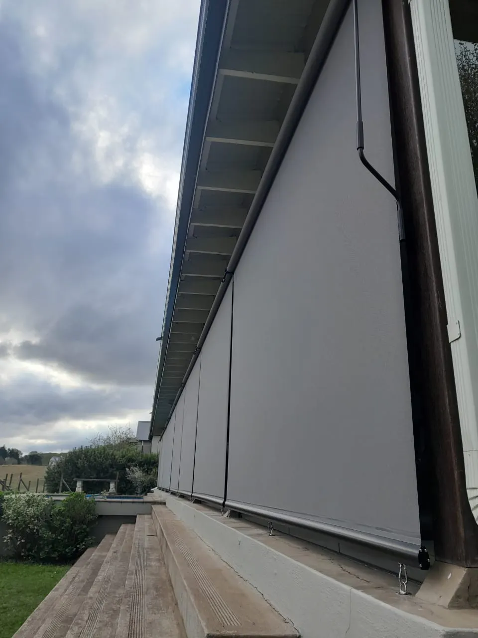 Exterior grey roller blinds lowered along a house veranda, shielding concrete steps and patio; metal fixings visible, with overcast sky and garden landscape
