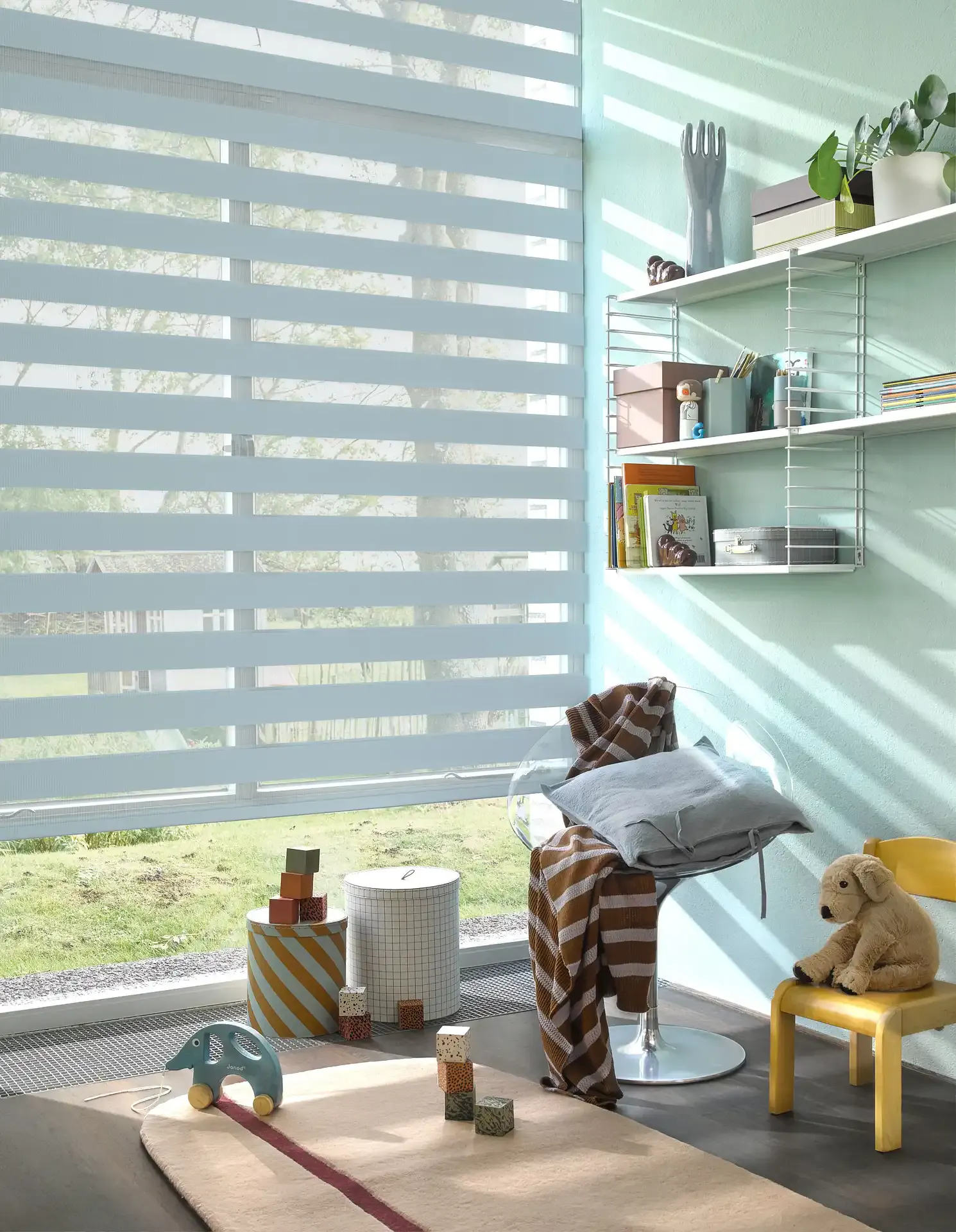 Double roller blind filters sunlight over a large window while a mint-painted kids' room displays wall shelves, toys, a striped throw on a clear chair and a