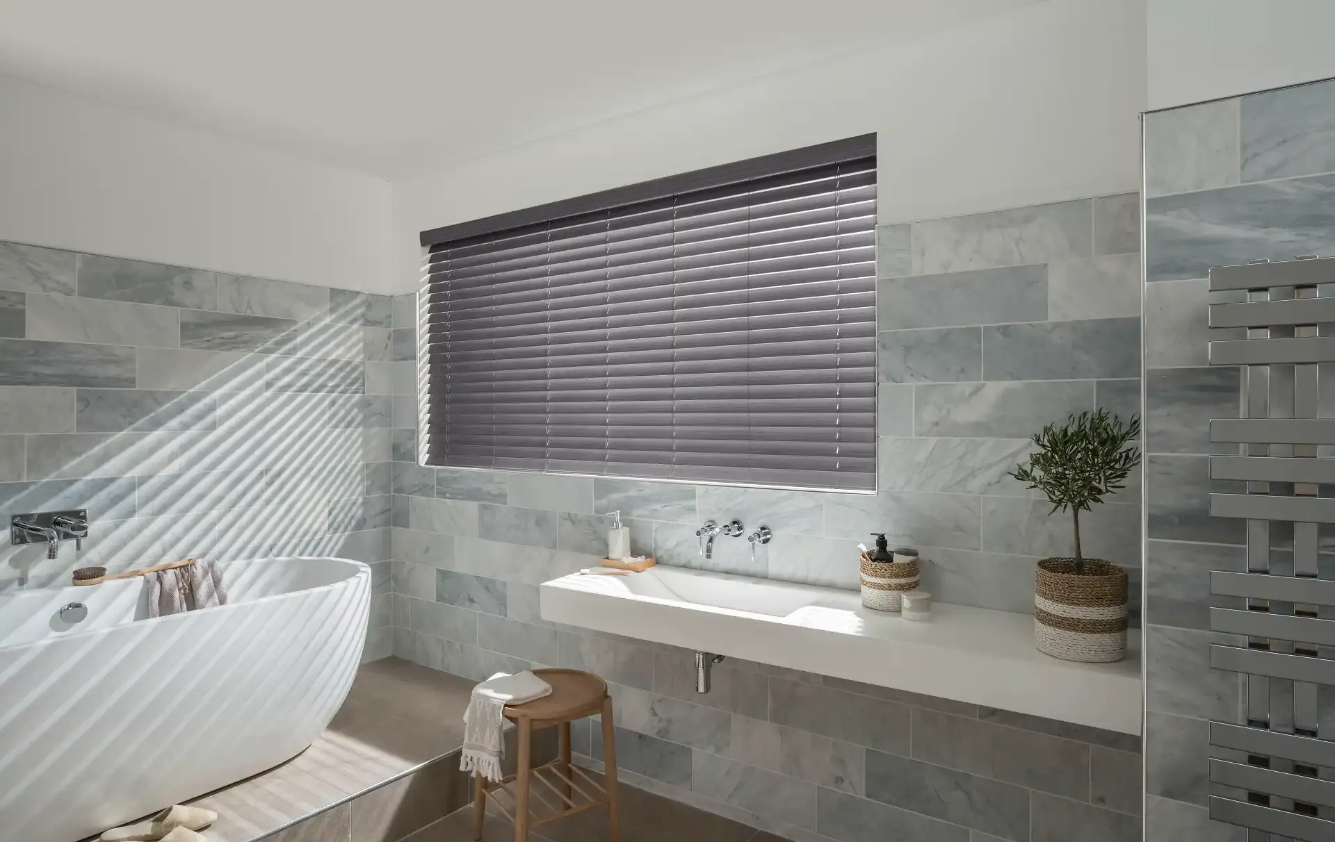 Grey horizontal window blind blocking light, casting striped shadows across a marble-tiled bathroom with freestanding tub, wall-mounted sink, wooden stool and