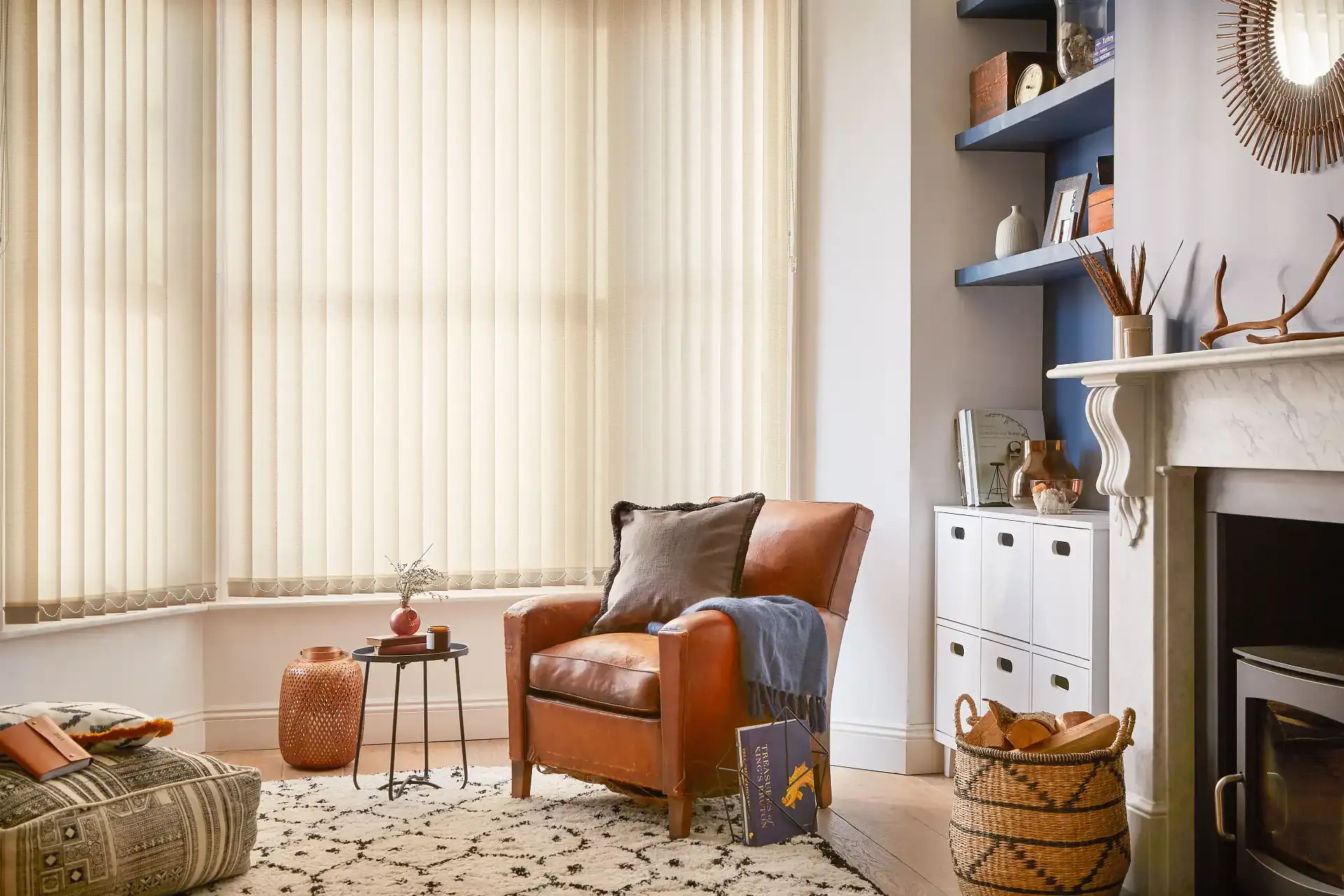 Vertical blinds filter soft daylight across a cosy living room with a leather armchair, side table, patterned rug, mantelpiece and built-in shelves with vases